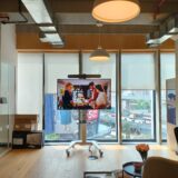 Modern office space featuring a portable video conferencing solution with an integrated display, camera, and soundbar on a trolley, positioned in front of large glass windows — ideal for hybrid meetings and dynamic collaboration.