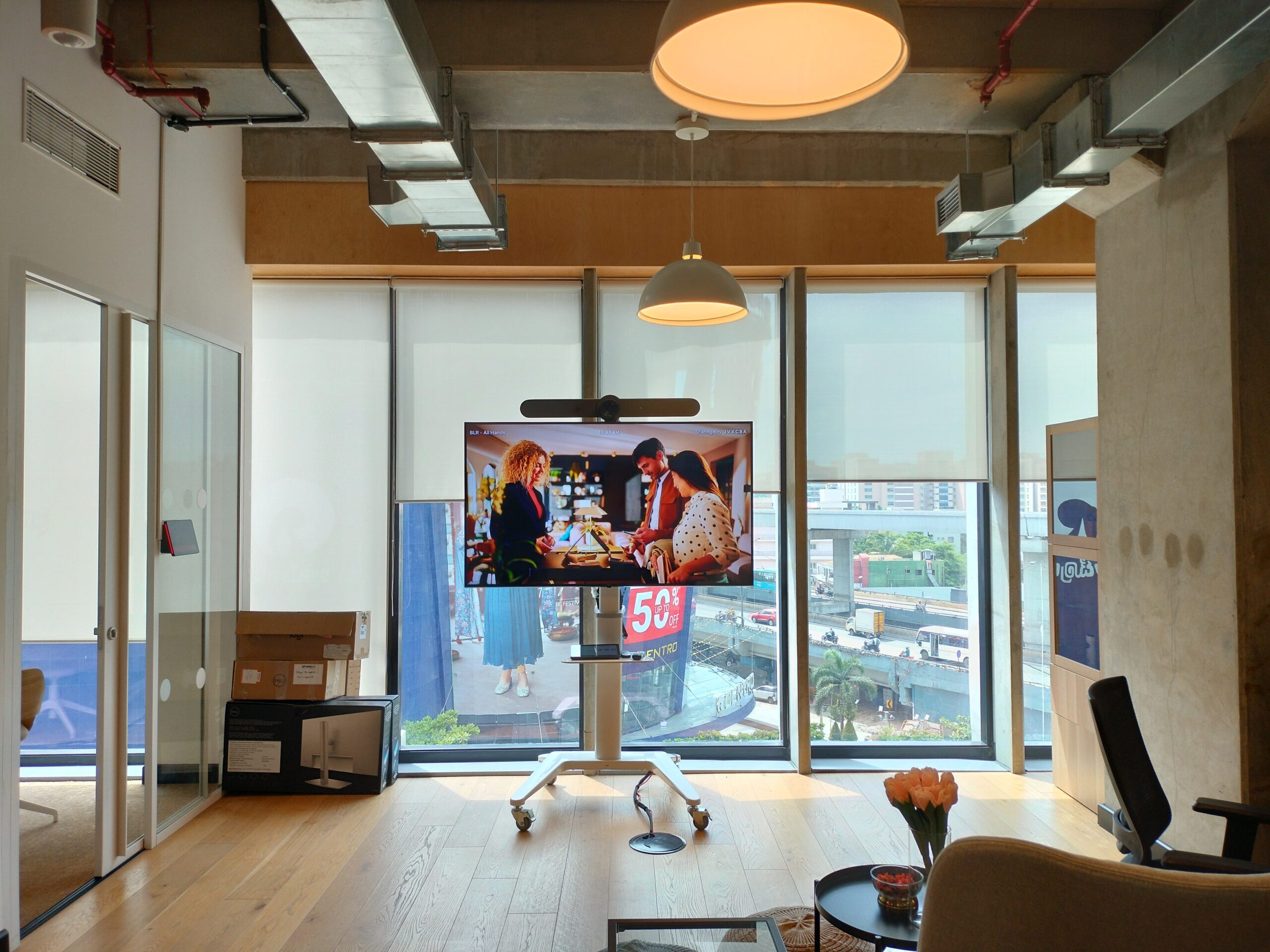 Modern office space featuring a portable video conferencing solution with an integrated display, camera, and soundbar on a trolley, positioned in front of large glass windows — ideal for hybrid meetings and dynamic collaboration.